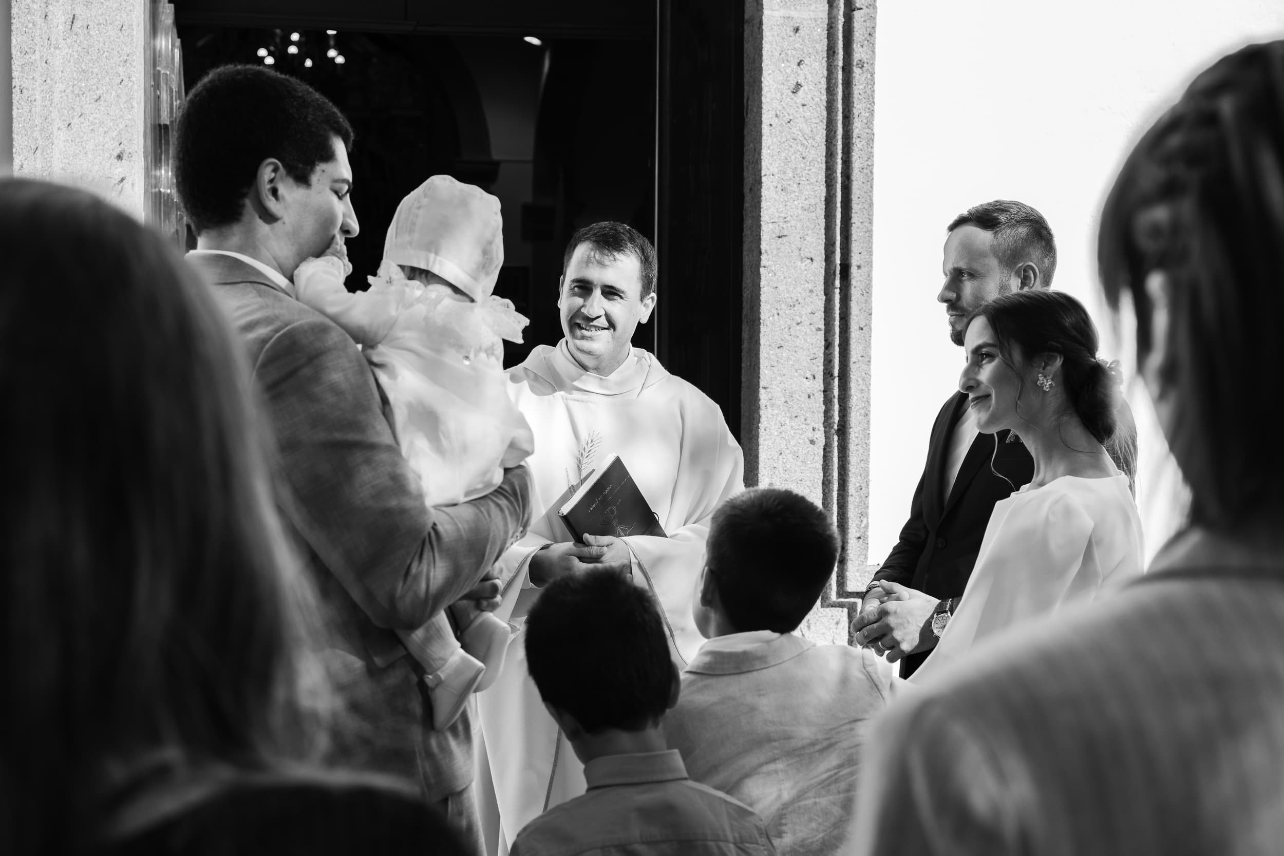 À porta da igreja, o sacerdote recebe a família com o bebé ao colo — momento espontâneo, a preto e branco